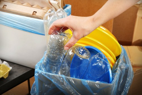 A close-up view of a person's hand placing a clear plastic water bottle into a large blue recycling bag. The bag already contains several other recyclable items, including yellow and blue plastic containers, and is situated inside an indoor environment, likely a commercial or warehouse space. The containers have a smooth, glossy surface, with some showing ridges or textured patterns typical of plastic packaging. The setting includes a black surface or table on the left, with a cardboard box or similar packaging visible in the background, hinting at waste sorting or recycling activities. The lighting is consistent and bright, highlighting the shiny plastic items and the person's hand. This scene aligns with waste collection and recycling services, such as those provided by Commercial Waste Canary Wharf, for managing plastic waste in the local business environment near Canary Wharf, London postcode E14.