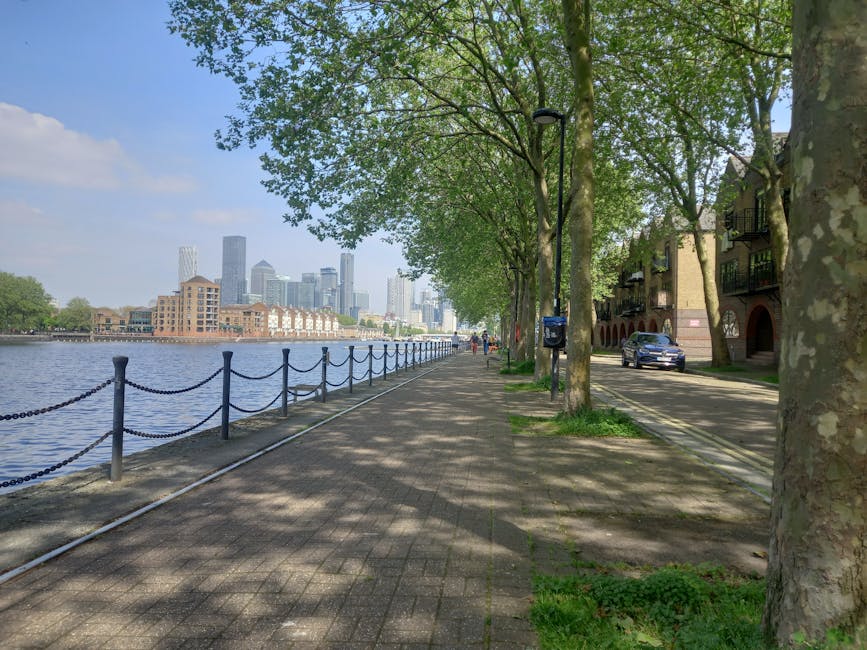 A tall, modern skyscraper with a predominantly glass exterior reflecting the sky and surrounding buildings stands prominently in this cityscape, positioned along a waterway with a smooth, dark surface