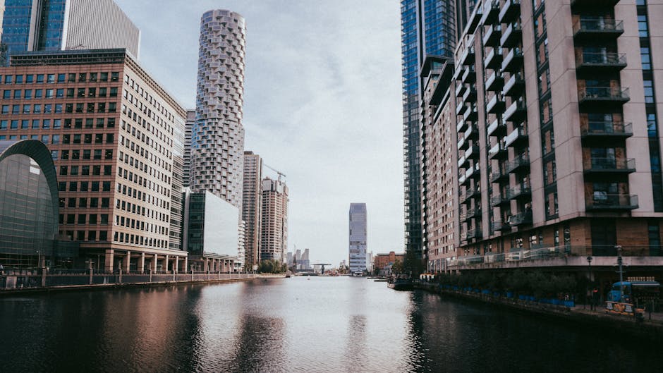 A cityscape featuring modern high-rise office buildings situated along a waterway, with the tallest building in the center having a sleek glass facade reflecting the sky and surrounding structures. To