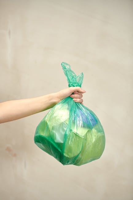 A person's hand holding a green and blue tinted plastic rubbish bag by its twisted knot top, with the bag appearing somewhat transparent and filled with waste. The bag is positioned horizontally acros