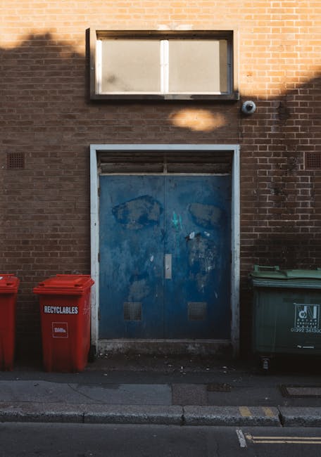 A person's hand holding a green and blue tinted plastic rubbish bag by its twisted knot top, with the bag appearing somewhat transparent and filled with waste. The bag is positioned horizontally acros