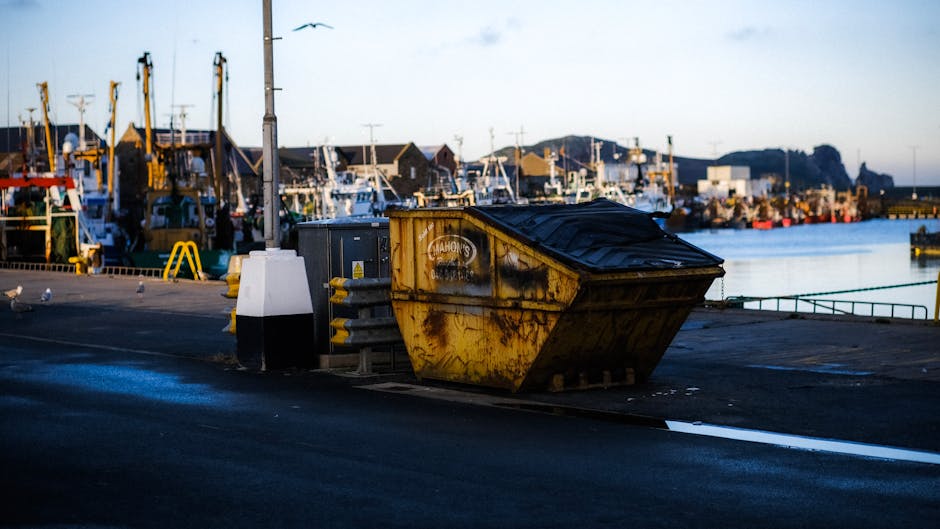 A worker wearing a face mask and safety gloves is standing on the edge of a large, open-top waste container, which is filled with various discarded furnishings and materials. The bulk of the rubbish i