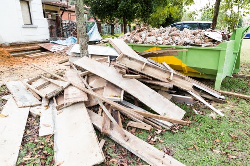 A pile of discarded wooden construction debris, including weathered and broken planks, visible on a patch of grass near a waste disposal site. The debris consists of aged, rough-surfaced wood with peeling paint and splintered edges, placed in front of a large green skip filled with bricks and rubble. In the background, there are trees with leafy branches, a paved driveway, and a building with white external walls, suggesting a residential or semi-urban setting. The scene appears to be part of a rubbish removal process, typical of a project in Canary Wharf or nearby London areas, managed by Commercial Waste Canary Wharf. The overall lighting is natural, with diffuse daylight illuminating the scene clearly, highlighting the textures and colors of the materials. The environment indicates an outdoor area where waste is temporarily accumulated for collection and disposal, fitting within the context of efficient waste solutions for local businesses or construction sites in the area.