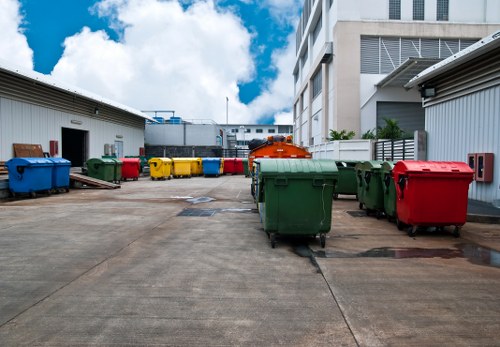 A paved outdoor yard area situated between industrial-style buildings, featuring multiple large wheelie bins in various colours including green, red, yellow, and blue, arranged in a row along the concrete surface. The bins appear to be made of durable plastic with a textured finish and are positioned with some slightly leaning or stacked, indicating recent use or movement. In the background, there are warehouse or service buildings with metal sidings, ventilation panels, and small access doors. The sky above is partly cloudy with patches of blue visible, casting natural daylight across the scene. The overall environment suggests a commercial rubbish collection or waste management area, consistent with services offered by Commercial Waste Canary Wharf for local businesses within the London docklands area, near Canary Wharf or Tower Hamlets. The scene captures a typical rubbish disposal zone with ample space for waste collection vehicles, highlighting the type of outdoor storage suitable for routine waste removal activities.