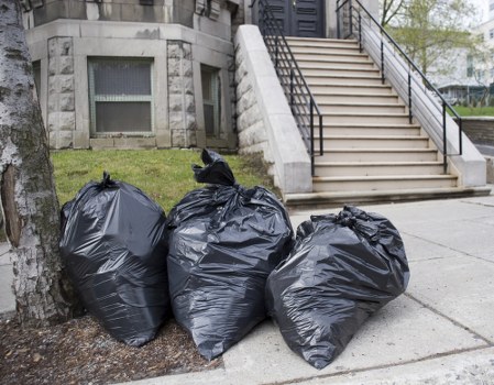 Three large black garbage bags filled with waste are stacked on a paved sidewalk near the base of a tree in an urban environment, with a staircase leading up to a building entrance visible in the background. The bags are made of shiny plastic material and are tied securely at the top, indicating they are ready for collection. The setting appears to be in a city area, possibly in Canary Wharf, with the pavement and stairs providing context for commercial waste disposal. The scene is well-lit, with natural daylight illuminating the bags and surroundings, and the environment suggests a routine rubbish removal process typically handled by services such as Commercial Waste Canary Wharf, which specialises in rubbish collection for local businesses.