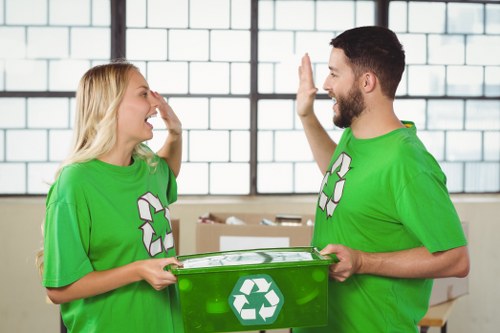 A photograph shows two young adults, a woman and a man, standing indoors in front of a large industrial-style window with a grid pattern. They are wearing bright green T-shirts with white recycling symbols on the front. The woman has long blonde hair and is smiling broadly while touching her nose with her right hand, and the man has short dark hair and a beard, smiling back and giving a high five with his right hand. The man is holding a green recycling bin with a white recycling logo on the side in front of him, which appears to be made of plastic and has a lid. The background includes a white brick wall and some cardboard boxes in a work or warehouse environment. The scene conveys a friendly and collaborative atmosphere related to waste management, highlighting the roles of Commercial Waste Canary Wharf in recycling and rubbish collection services within the local area near Canary Wharf.